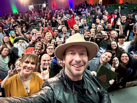 A man in a leather jacket and tan hat takes a selfie with a large and enthusiastic group of people behind him in an auditorium setting. The diverse crowd, many of whom are smiling and making playful gestures like peace signs, fills the space. A woman in a mustard-colored blouse, wearing a necklace, stands out in the foreground, also smiling widely. The atmosphere seems lively, filled with excitement and camaraderie.