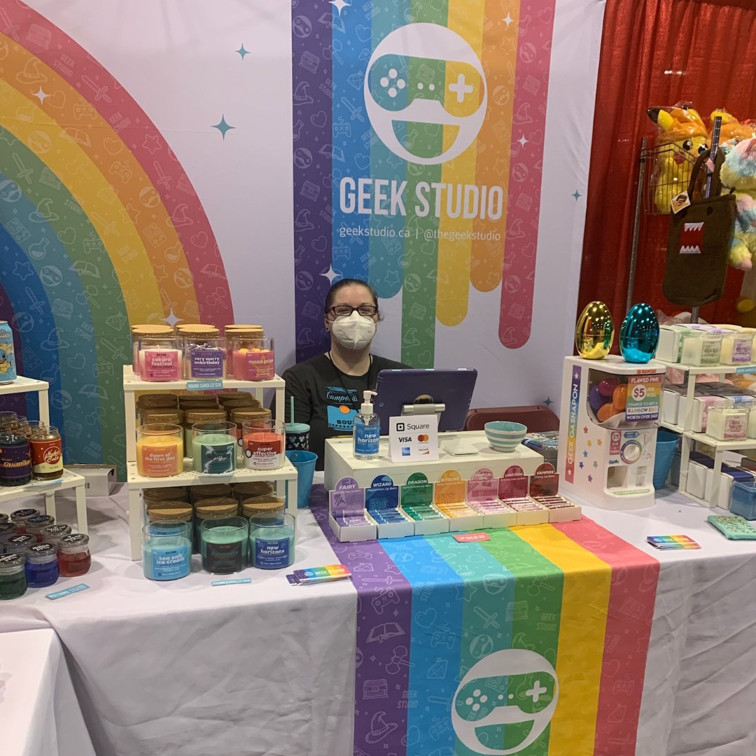 A woman is seated in front of a table. She has a table with products in front of her and a sign behind her that reads "Geek Studio"