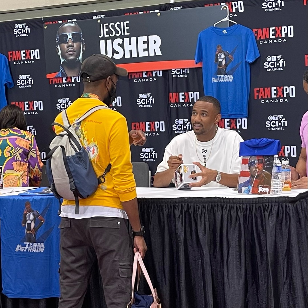 Jessie Usher is seated in front of a table. He is wearing a white t-shirt and he is signing a Funko Pop. He is speaking to a fan while signing the Funko pop. The fan is wearing black pants, a yellow sweater and a black hat.