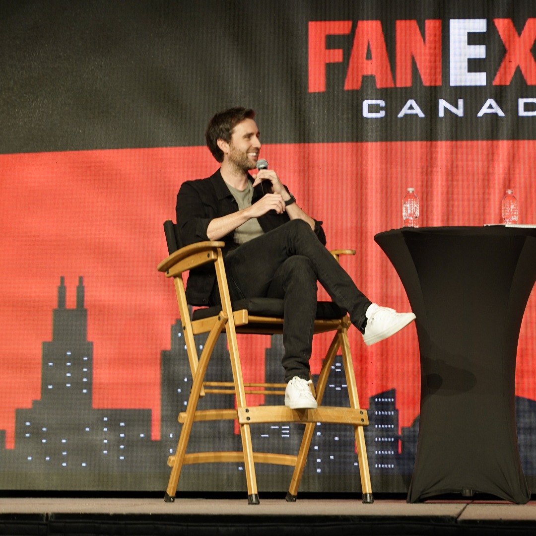 Matthew Lewis is wearing a black blazer with a green shirt underneath and black jeans. He is sitting on stage at FAN EXPO Canada for his panel.