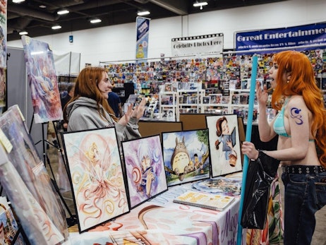 Two women are in a convention setting filled with various artwork displays. On the left, a woman with long reddish-brown hair, wearing a grey hoodie, animatedly talks while gesturing with her hands. She stands behind a table showcasing vibrant art prints, including fantasy-themed characters. On the right, a woman with bright orange hair, styled in a cosplay outfit, listens intently. She has a blue swirl tattoo on her shoulder and holds a light blue prop. Behind them, the convention continues with various stalls, including one labeled "Great Entertainment." The ambiance is lively, filled with creativity and fandom discussions.