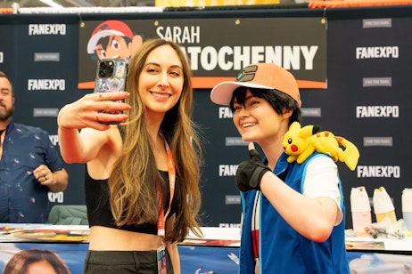 The image captures a cheerful moment at FAN EXPO, showing Sarah Natochenny and a female fan posing for a selfie. On the left, Sarah with long dark hair, wearing a black crop top and smiling broadly, holds up her phone to take a photo. On the right, another woman dressed as Ash Ketchum from Pokémon, complete with a baseball cap, blue vest, and a Pikachu plush on her shoulder, poses enthusiastically with a thumbs-up. They are in front of a backdrop that includes the text "FAN EXPO" and "SARAH NATOCHENNY," indicating the context of the event.