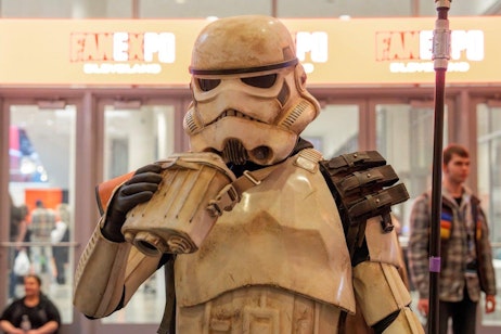 The image depicts a person dressed as a stormtrooper from "Star Wars" at a FAN EXPO event, holding up a canister to its helmet as if drinking. The costume is detailed and weathered, suggesting battle-worn armor. Behind the stormtrooper, the brightly lit FAN EXPO Cleveland sign is visible, indicating the event's location. The setting includes other attendees and displays typical of such conventions, creating a lively atmosphere.