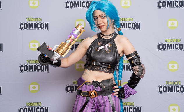 A Jinx (Arcane) cosplayer poses with her prop blaster. She holds the blaster facing upwards and one hand on her hip.