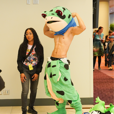 A young woman in a dragon ball z tshirt poses with with a half frog cosplayer with a cartoonish frog head.