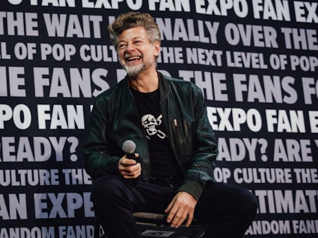 Andy Serkis with salt-and-pepper hair and beard sits holding a microphone, wearing a dark green jacket with a black t-shirt that has a skull graphic. He is laughing against a bold black and white backdrop filled with repeated phrases like "FAN EXPO," "THE NEXT LEVEL OF POP CULTURE," and "ARE YOU READY?" The ambiance suggests an engaging fan convention or interview setting.