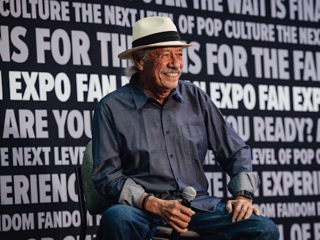 Edward James Olmos with a warm smile, wearing a white hat and a blue-gray shirt, sits holding a microphone. He is against a bold black and white backdrop with phrases like "FAN EXPO," "THE NEXT LEVEL OF POP CULTURE," and "ARE YOU READY?" suggesting a lively convention or interview scene.
