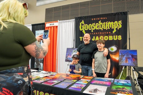 The image captures a heartwarming moment at a fan convention booth dedicated to "Goosebumps" illustrator Tim Jacobus. A woman, with her back to the camera, is taking a photo of two young boys with Tim, all smiling in front of the booth. The booth features a vibrant display of colorful artwork, including paintings and character illustrations from "Goosebumps." The backdrop proudly displays the "Goosebumps" logo alongside the name 'Tim Jacobus', enhancing the theme of the booth. The setting is busy with other attendees and exhibits in the background, contributing to the lively atmosphere of the event.
