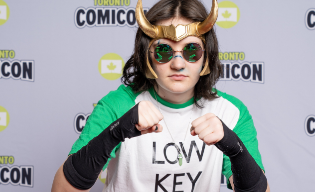 A cosplayer dressed as Low Key Loki, wearing a white and green baseball shirt, green sunglasses, and Loki horns.