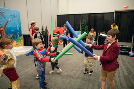 The image features a playful scene at a children's activity area during a fan convention. Several children are engaged in a spirited battle using foam swords and pool noodles, each child donned in different costumes, including Spider-Man and martial arts attire. Their expressions are filled with joy and excitement as they playfully duel each other. In the background, other children and adults can be seen participating in or watching the activities, contributing to a lively and festive atmosphere. The setting includes a vibrant backdrop with a marine theme, enhancing the playful and imaginative environment.