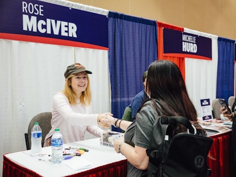 Rose McIver, a cheerful woman wearing a hat, sits at a signing table, extending her hand to greet a fan. The table is adorned with markers, bottled water, and promotional materials. To the right, another booth banner reads "Michelle Hurd." A fan, seen from behind, stands opposite McIver, holding a mobile phone and wearing a backpack. The scene suggests a fan convention or autograph session.