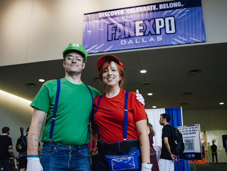 A man (left) dressed as Luigi from Mario Bros, and a woman (right) dressed as Mario, with the FAN EXPO Dallas banner in the background.