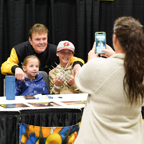Cal Dodd poses with two young fans as their mother takes a photo. Cal wears a black and yellow X-Men jacket, the little girl is in a blue rain jacket and her brother is in a beige hurley hoodie with wolverine claws. Mom is in a white cardigan and holds up an iphone to take a photo