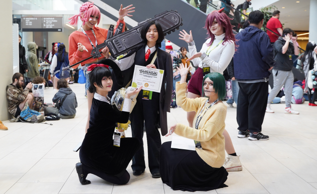 Snacck Box pose with one of their cosplay hallway award winners, they are holding a large chainsaw/sword hybrid prop