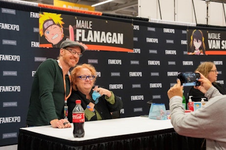 The image captures a heartwarming moment at a FAN EXPO event where a Chris Edgerly and Maile Flanagan are smiling and posing together for a photograph at an autograph table. Behind them is a banner featuring the name "Maile Flanagan" with a cartoon image of Naruto Uzumaki. Maile is making a Naruto gesture with her hands, while Chris leans in close with a smile. A third person is seen taking their photo. This scene conveys the friendly and engaging atmosphere typical of fan conventions.