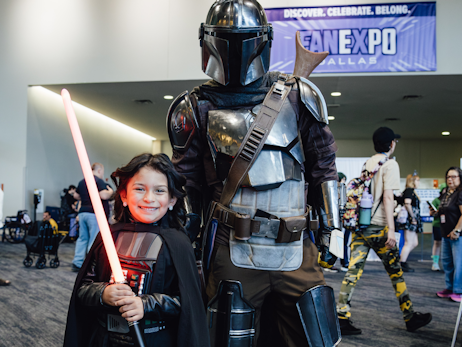 A young girl (left), smiling and dressed as Darth Vader, holding a red lightsaber. An adult (right) cosplaying as a Mandalorian.