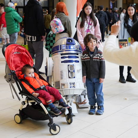 A mother takes a photo of her two kids with R2-D2. One child, in various shares of red, is in a stroller, the other smiles, wearing a blue caridgan and blue jeans.
