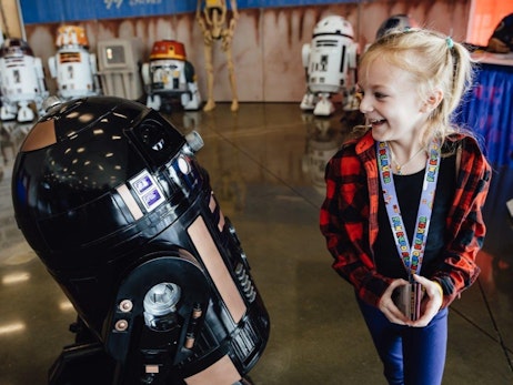 A delighted young girl with blonde hair tied in a ponytail, wearing a red plaid shirt and a lanyard, interacts with a large black droid. In the background, several other Star Wars-themed droids and a banner are visible, suggesting the setting is a fan convention or themed event. The girl's expression is one of wonder and amusement as she gazes at the droid.
