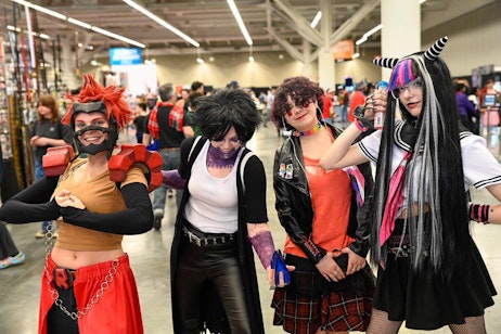 The image features four individuals in elaborate and colorful costumes posing confidently at a fan expo. From left to right: the first person is dressed as a character with spiky red hair and oversized gauntlets, showcasing a big smile and an animated pose; the second person is styled with black hair and a punk rock look, making a playful gesture; the third person, in red and black with a punk aesthetic, strikes a confident pose; the fourth person is costumed in a gothic Lolita outfit with long, black and pink hair, holding a small bottle, and making a peace sign. The backdrop of the expo includes other attendees and booths, creating a vibrant and festive environment typical of such events.
