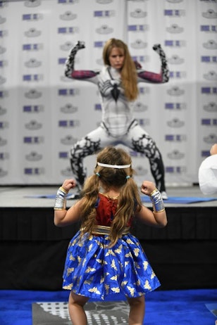 A little girl, dressed up like Wonder Woman, with her back to the camera - has her arms up showing strength as her joins in on superhero yoga