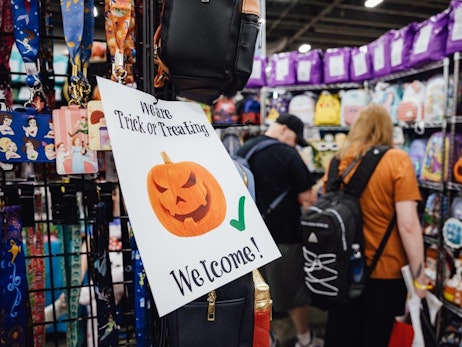 A festive sign reading "We are Trick or Treating - Welcome!" with an illustration of a smiling jack-o'-lantern is prominently displayed in a convention booth. The booth showcases an array of merchandise, including colorful lanyards, wallets featuring animated characters, and bags hanging on display racks. In the blurred background, attendees, one with an orange shirt and black backpack, peruse the items for sale. The atmosphere suggests a fun and lively event with a Halloween theme.