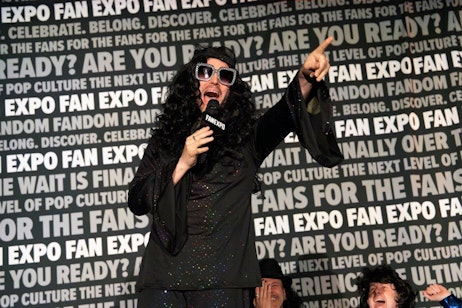 The image captures Elijah Wood at FAN EXPO, dressed in a flowing black shirt and sporting long, curly black hair, complete with sunglasses. He is dynamically posed, singing into a microphone while pointing upwards with his other hand, expressing excitement. His expressive face and lively gesture convey a strong sense of enthusiasm and engagement with the audience. In the background, the repeating text "FAN EXPO," along with various slogans about pop culture and fandom, enhances the festive and energetic atmosphere of the event.