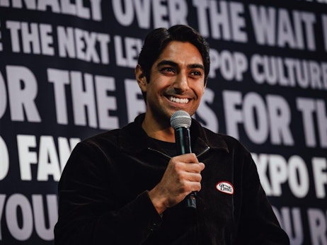 Eman Esfandi with dark hair and prominent features holds a microphone, speaking enthusiastically during an event. He's dressed in a dark-colored zip-up sweater with a "Bruh" badge pinned on it. Behind him, a large backdrop with bold, partially visible text alludes to a pop culture or fan-related event. The man's genuine smile and engaging demeanor suggest he's addressing an attentive audience.