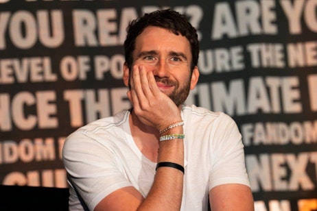 The image features Matthew Lewis sitting in front of a backdrop with "FAN EXPO" logos and text about pop culture. He wears a casual white V-neck t-shirt and multiple bracelets. His expression is playful and thoughtful as he smiles slightly, resting his chin on his hand. His gaze seems directed towards someone or something off-camera, contributing to a relaxed and engaging atmosphere. The setting suggests he is a guest or speaker at the event, interacting with attendees or perhaps amid a discussion or interview.