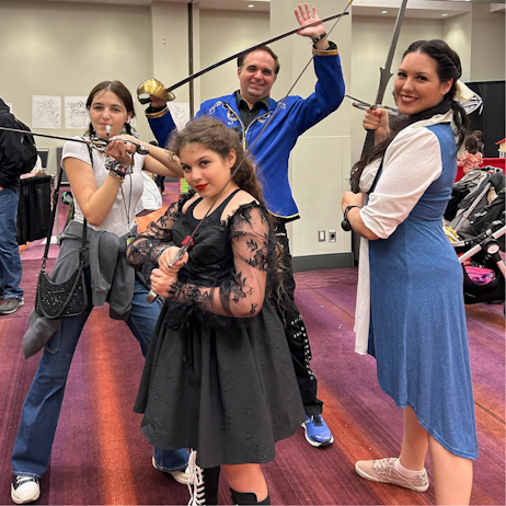 Smiling for the camera, a family holds the swords of the oxford ren festival. Left to right, the oldest daughter is not in cosplay, front centre, youngest daughter is Wednesday Addams. Behind her, dad is Adam from Beauty and the beast, and right, mom is Belle from Beauty and the Beast