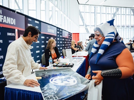 At a bustling fan expo event, Eman Esfandi in a white shirt stands behind a table adorned with merchandise, flashing a genuine smile as he interacts with a woman dressed in an elaborate costume. The woman, in blue and white face paint and a headpiece, seems thrilled as she shows a poster to the man. Behind them, event banners display names, including "FANEXPO" and other unidentified individuals engage in similar interactions, making the setting vibrant and lively.
