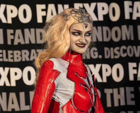 The image features a person in an elaborate costume at FAN EXPO. They are dressed as fantasy or horror character with striking makeup and detailed costuming. The costume includes a shiny red bodysuit with metallic armor accents and a crown-like headpiece adorned with ornate designs. The makeup is dramatic, with pale skin and dark, exaggerated features that enhance the character's intense, menacing expression. The background has a black banner with white text repeating "FAN EXPO" and phrases celebrating fandom and pop culture, suggesting a vibrant and engaging event atmosphere.