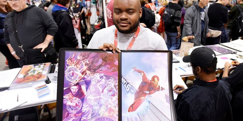 Jamal Campbell in a light grey sweater and a red lanyard holds up original art featuring Doctor Strange and Iron Man