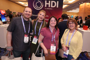 Four IT professionals smiling together in the Expo Hall in front of the HDI booth, representing the networking, community, and career growth opportunities at HDI Service & Support World.