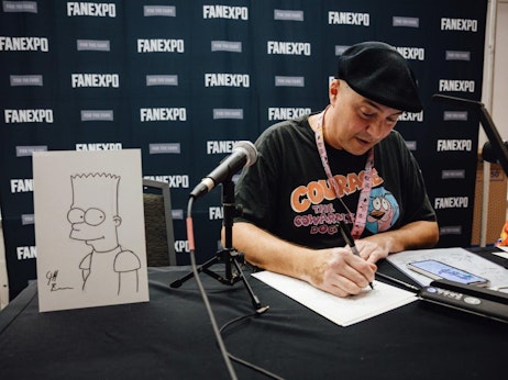 A man wearing a black hat and a t-shirt with "Courage the Cowardly Dog" graphics sits at a table, sketching on a paper. Next to him stands a drawing of the cartoon character Bart Simpson on a white board. The backdrop features repeated "FANEXPO" logos, suggesting the setting is a fan convention. There's a microphone on the table, and the man is surrounded by various drawing tools and a digital device displaying artwork.