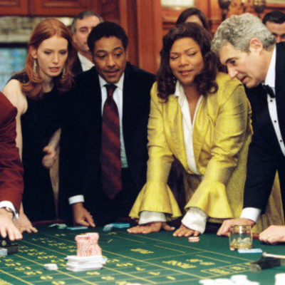 Giancarlo in a suit next to Queen Latifah in a yellow dress at a roulette table, surrounded by people.