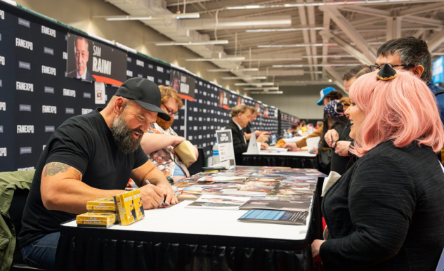 The image showcases a lively and engaging moment at a FAN EXPO event, featuring Ethan Suplee signing autographs. He's wearing a black t-shirt and cap and is laughing heartily as he interacts with fans. One of the fans, a woman with pink hair, eagerly watches him sign, appearing delighted by the interaction. The background is bustling with other attendees and artists at their booths, with promotional banners, including one that reads "Sam Raimi," adding to the fan-centric environment of the expo.
