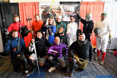 The image features a diverse group of individuals dressed in various fencing and medieval combat gear, posing together at a FAN EXPO event. The group, consisting of men and women, displays a range of costumes, from traditional fencing whites to colorful outfits that resemble characters from fantasy realms. Some wield swords, other lightsabers, and even medieval weapons, highlighting the blend of historical and fantastical themes. The backdrop includes a banner for "Two Ravens Fencing School," indicating their affiliation or the event they are promoting. This engaging scene captures the unique intersection of sport and fandom culture, providing an interactive experience for attendees.