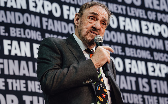 John Rhys-Davies with a distinctive expression speaks into a microphone. He's dressed in a dark suit paired with a colorful tie adorned with various small emblems. He stands in front of a backdrop with repeated fan expo text, indicating he's part of a panel or event.