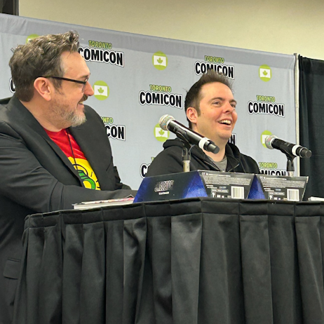 Tim Sheridan (far left, glasses, greying facial hair, red shirt, black blazer) and Jeremy Adams (right, grey shirt, black hoodie) with microphones in front of them, smile as they discuss their work on Green Lantern
