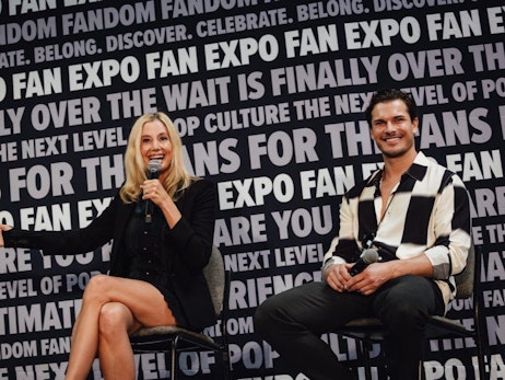 At a FAN EXPO event, Mira Sorvino energetically speaks into a microphone while gesturing with her free hand. She's seated and wearing a black blazer over a dark dress. Beside her, Gleb Savchenko in a black and white striped shirt sits relaxed, holding a microphone and smiling towards the audience. The backdrop is filled with repeated "FAN EXPO" text and various other slogans, creating an immersive atmosphere for the event.