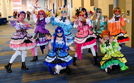 The image captures a group of eight individuals in vibrant and elaborate cosplay outfits, striking playful poses at a convention. They are dressed in colorful maid costumes with varying shades of pink, purple, blue, green, orange, and yellow, each adorned with frills, bows, and unique accessories. Their wigs are equally striking, ranging in hues that match their outfits. The setting suggests a fun and energetic atmosphere typical of a FAN EXPO event, with each participant expressing joy and character through their poses and expressions.