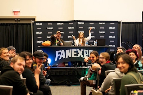 The image shows an engaging and joyful scene at a FAN EXPO panel discussion. Two panelists, a man in a hat and voice actor Sarah Natochenny, are seated behind a table adorned with the FAN EXPO logo, enthusiastically interacting with the audience. The man is pointing into the crowd while Sarah gestures with both arms raised, sharing a laugh with attendees. The audience in the foreground is diverse and lively, some posing for the camera with smiles and playful hand gestures. The overall atmosphere is vibrant and participatory, typical of a lively panel session at a fan convention.