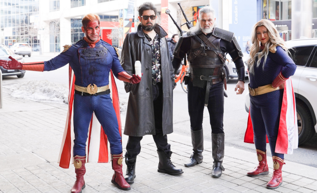Four cosplayers stand on the sidewalk outside of the convention centre - from left to right: Male Homelander, Billy Butcher, Geralt of Rivea, Female Homelander