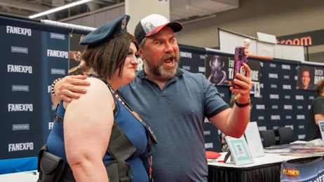 The image captures a joyful and humorous moment at the FAN EXPO. Roger Clark, wearing a blue polo shirt and a trucker cap animatedly takes a selfie with a young woman in a police-themed cosplay costume, featuring a cap and a sleeveless top. The woman, smiling broadly, hugs the man from the side. The background displays the FAN EXPO logo and various celebrity signs, reflecting the vibrant atmosphere of the event. Roger's expression and posture add a playful vibe to the scene, highlighting the fun and community spirit typical at such fan conventions.