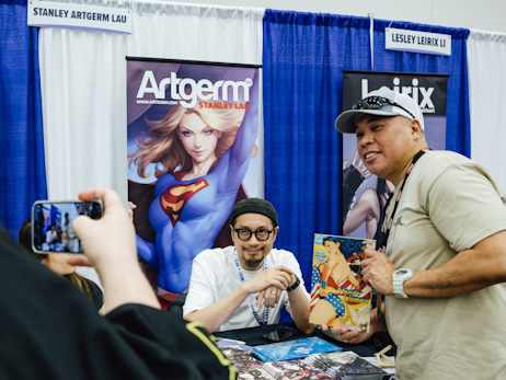 Stanley Artgerm Lau, sitting at his table, smiling and posing with a fan to his right for a photo.