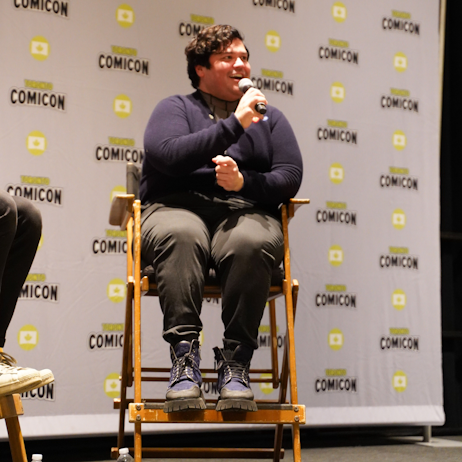 Harvey Guillen sits facing the audience, smiling as he answers a fan question. He is wearing black pants and a navy blue cardigan