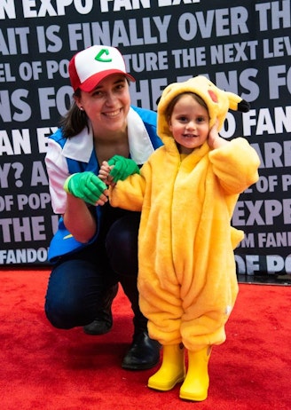 This image features a delightful scene at FAN EXPO with a woman and a toddler posing in front of a backdrop printed with the words "FAN EXPO." The woman, dressed in a "C" cap and a costume resembling Ash Ketchum from Pokémon, kneels next to the toddler, who is adorably dressed in a fluffy yellow Pikachu costume complete with bright yellow boots. Both are smiling joyfully, adding to the lighthearted, family-friendly atmosphere of the event. The vibrant red carpet and event-themed backdrop enhance the celebratory vibe.