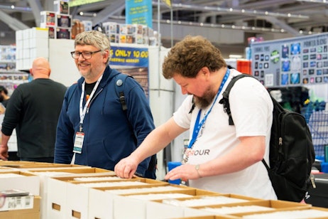 This image captures two men browsing through boxes of comics at a busy convention center. The man on the left, wearing glasses and a blue zip-up hoodie, is smiling and looking towards the camera, while the man on the right, dressed in a white T-shirt and carrying a backpack, is focused on carefully selecting comics. The setting is vibrant and filled with various stalls displaying pop culture merchandise, creating a lively atmosphere typical of a FAN EXPO event.