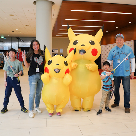 A family poses for a photo. Left to right: son, holding a minecraft sword, mom holds up a peace sign. Two children dressed as pikachu, one taller than the other. Right, another brother dressed as link, holds up her sword, and dad smiles for the camera