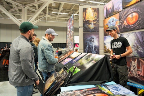 This image features two men browsing through a selection of vibrant and colorful artwork at a busy FAN EXPO event. They are standing in front of a stall adorned with large, eye-catching paintings on the walls, displaying various fantasy and science fiction themes. The first man, wearing a green cap and a grey sweater, is examining a piece intently. The second, in a denim jacket and glasses, is smiling as he looks through the artwork. The vendor, in a black Star Wars T-shirt and a cap, seems amused by something he is holding. The scene captures the lively, engaging atmosphere of the expo.